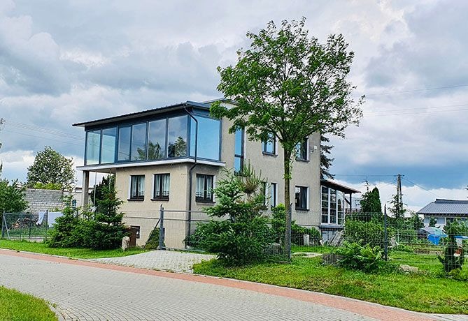 Dwukondygnacyjny dom z przeszkloną nadbudową na drugim piętrze, widok z zewnątrz, na pierwszym planie drzewo i chodnik z kostki.