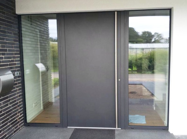 Nowoczesne, antracytowe drzwi wejściowe z aluminiową ramą oraz bocznymi panelami szklanymi, widok z zewnątrz na tle ceglanej ściany i zieleni.