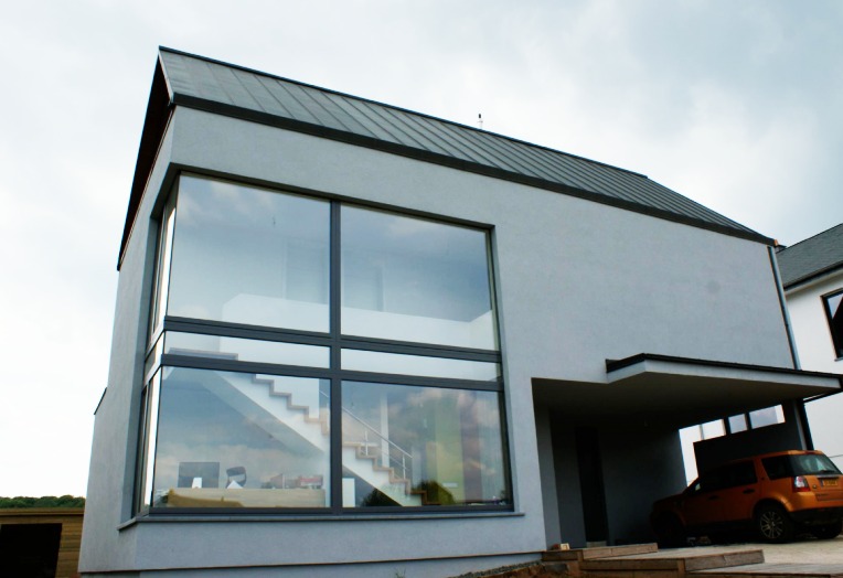 Nowoczesny dom z dużym oknem z czarnymi ramami, w którym odbija się wnętrze z białymi schodami, obok znajduje się wiata garażowa z pomarańczowym samochodem.