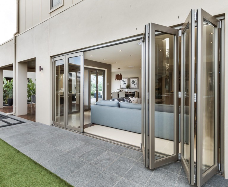 Składane, przeszklone drzwi balkonowe z aluminiową ramą, otwarte na taras wyłożony szarą kostką, widok na salon z sofą w tle.