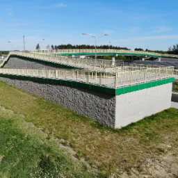 Betonowy wiadukt dla pieszych i rowerzystów z rampami, zielonymi akcentami i szarym kamiennym wykończeniem, wkomponowany w krajobraz z drogą i drzewami w tle.