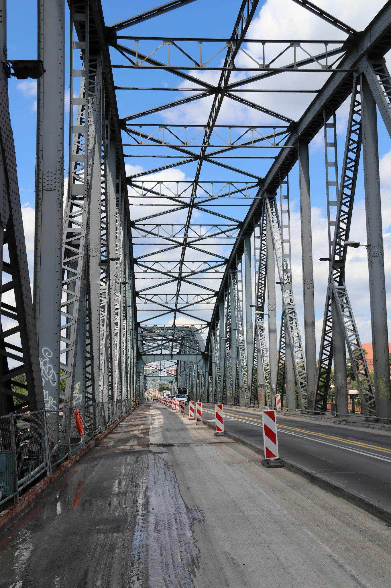 Stalowa konstrukcja mostu kratownicowego w trakcie remontu nawierzchni, widoczne bariery ochronne z czerwono-białymi pasami i tymczasowe ślady po wodzie.