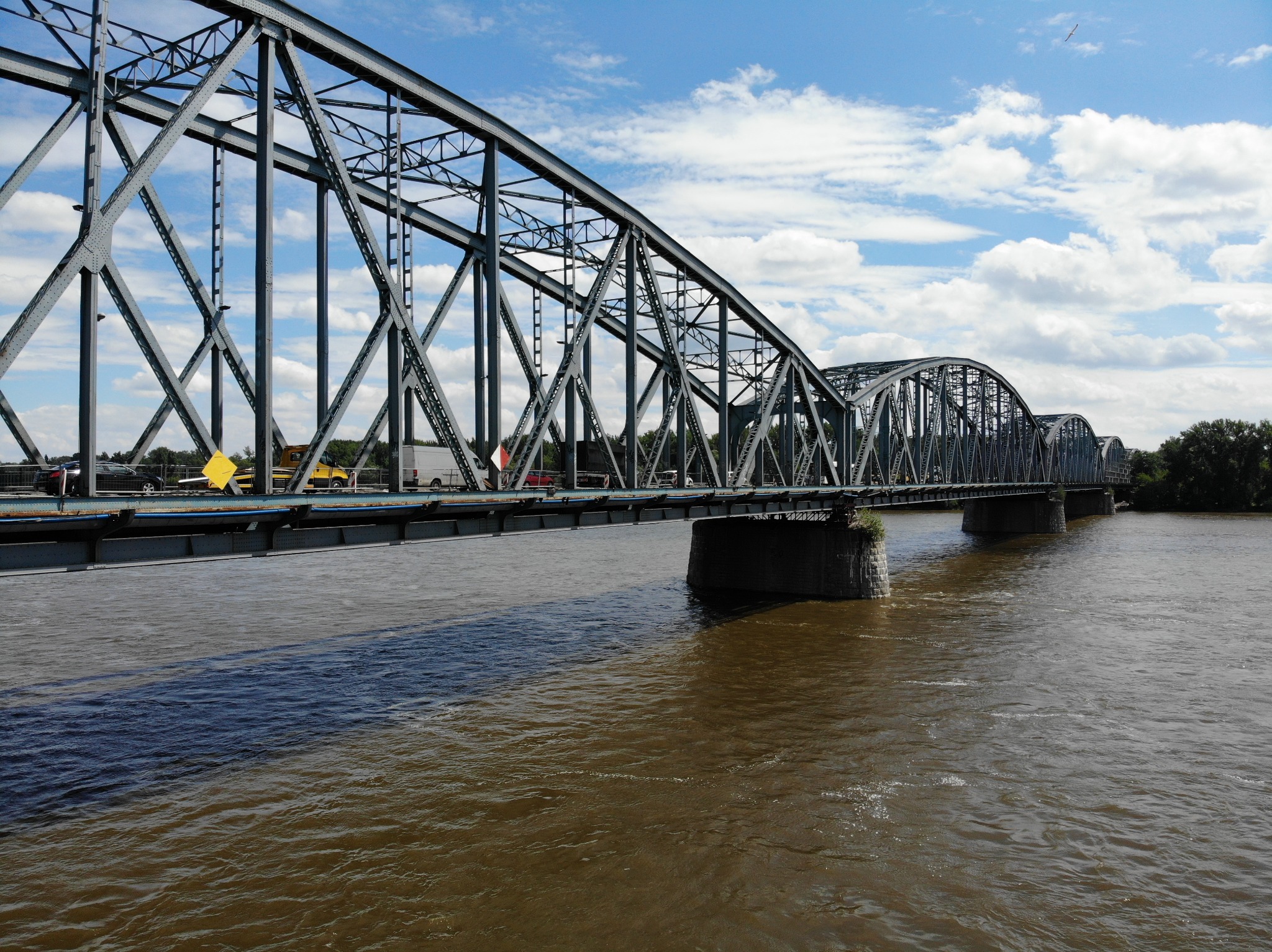 Stalowy most kratownicowy nad rzeką z pojazdami na nim, widok z dołu na konstrukcję i wodę.