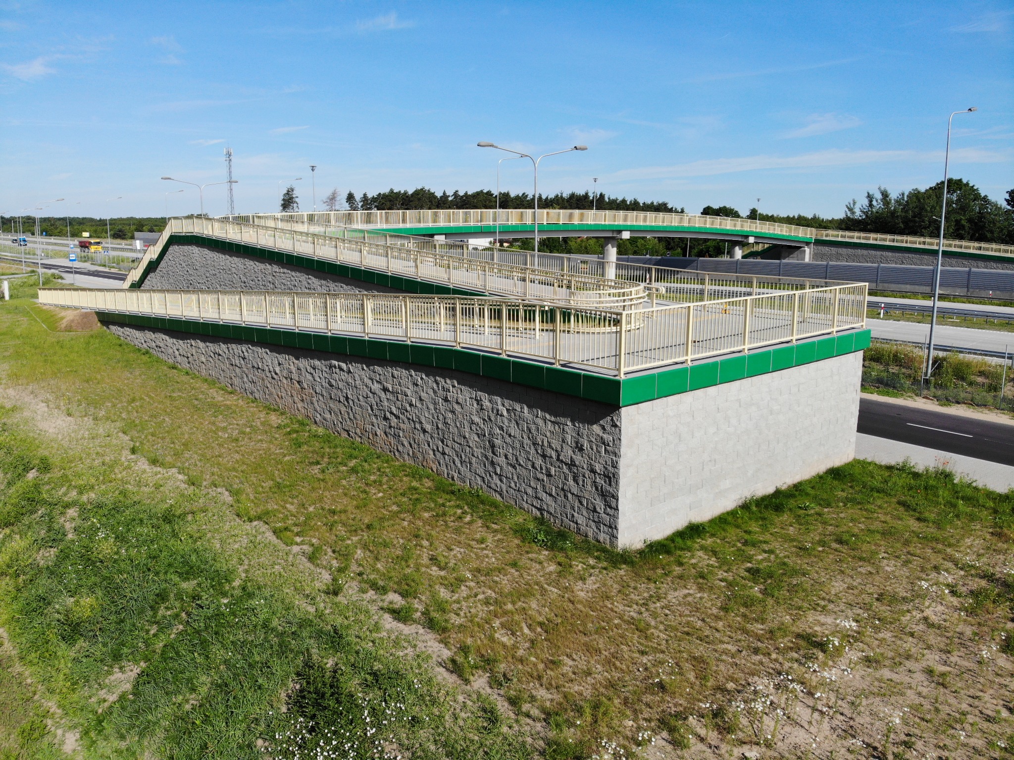 Betonowy wiadukt dla pieszych i rowerzystów z rampami, zielonymi akcentami i szarym kamiennym wykończeniem, wkomponowany w krajobraz z drogą i drzewami w tle.