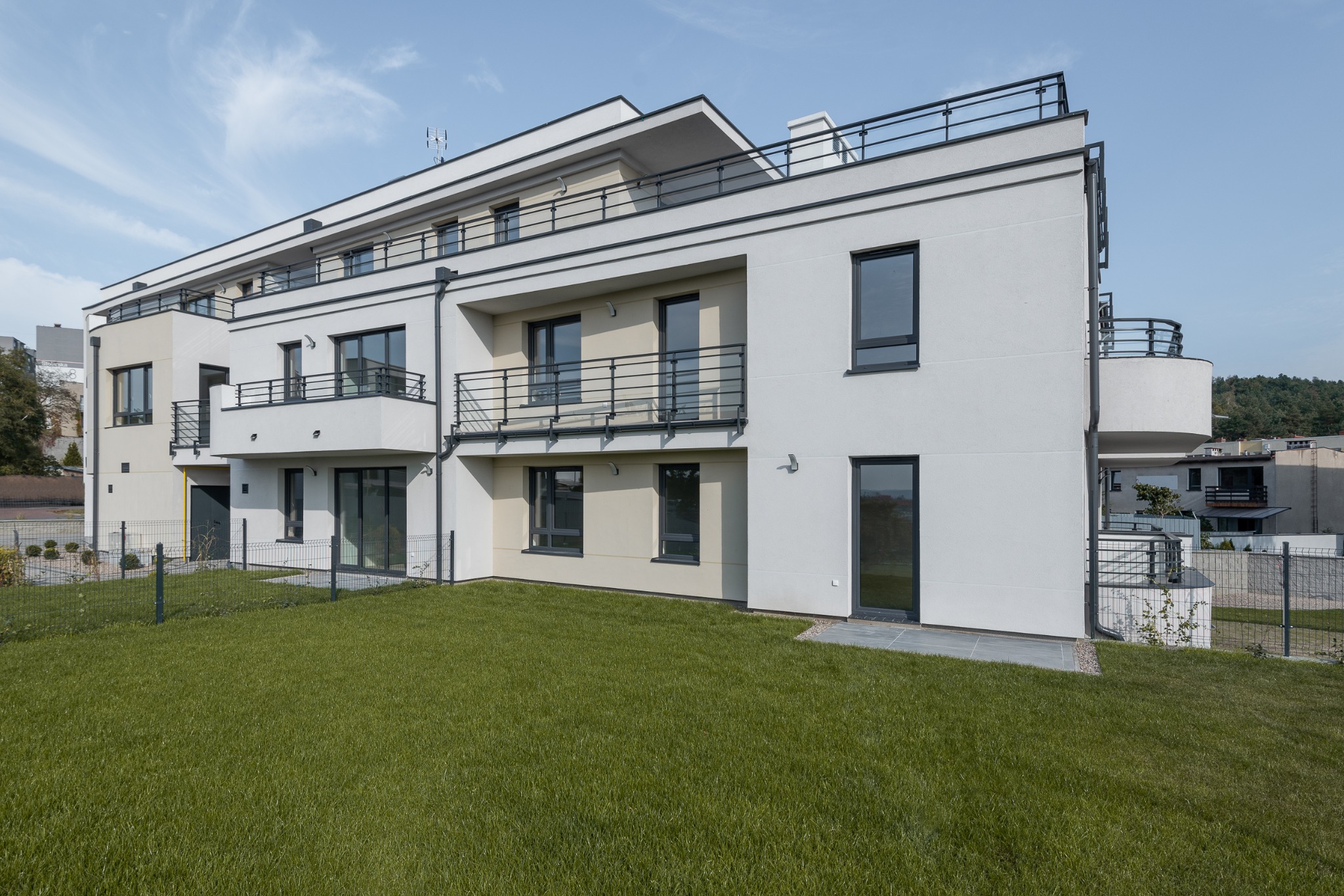 Nowoczesny, trzypiętrowy budynek mieszkalny z balkonami i płaskim dachem, otoczony trawnikiem i ogrodzeniem panelowym.