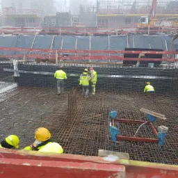 Ekipa budowlana w jaskrawych kamizelkach pracuje nad konstrukcją z siatki stalowej na placu budowy w mglisty dzień, widoczne szalunki i dźwig w tle.