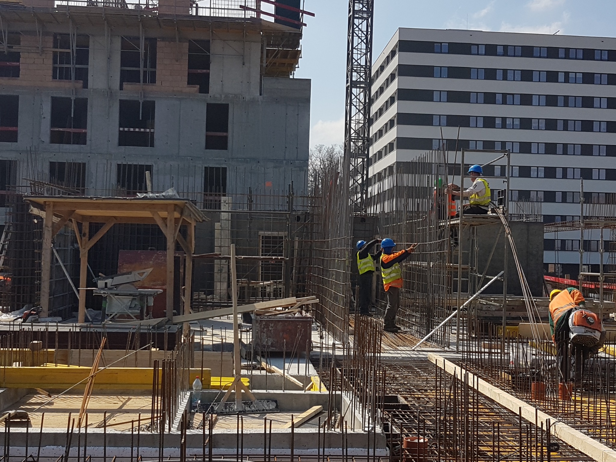 Wielopiętrowy budynek w surowym stanie z widoczną konstrukcją żelbetową, pracownicy w kaskach i kamizelkach odblaskowych montują zbrojenie, w tle drugi budynek i dźwig.