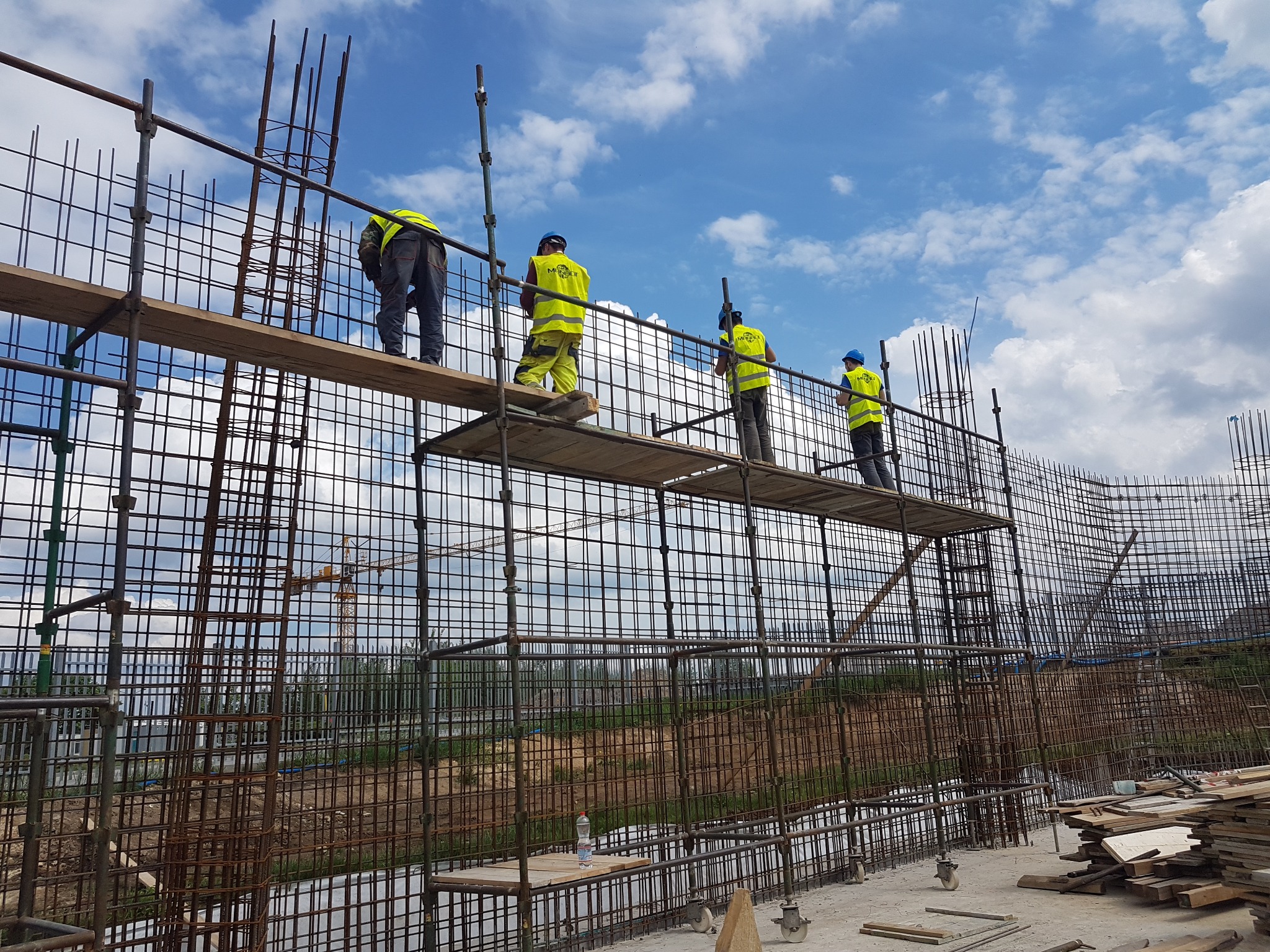 Czterech pracowników w żółtych kamizelkach i niebieskich kaskach pracuje na rusztowaniu przy zbrojeniu ścian budynku, widoczne niebo z chmurami.