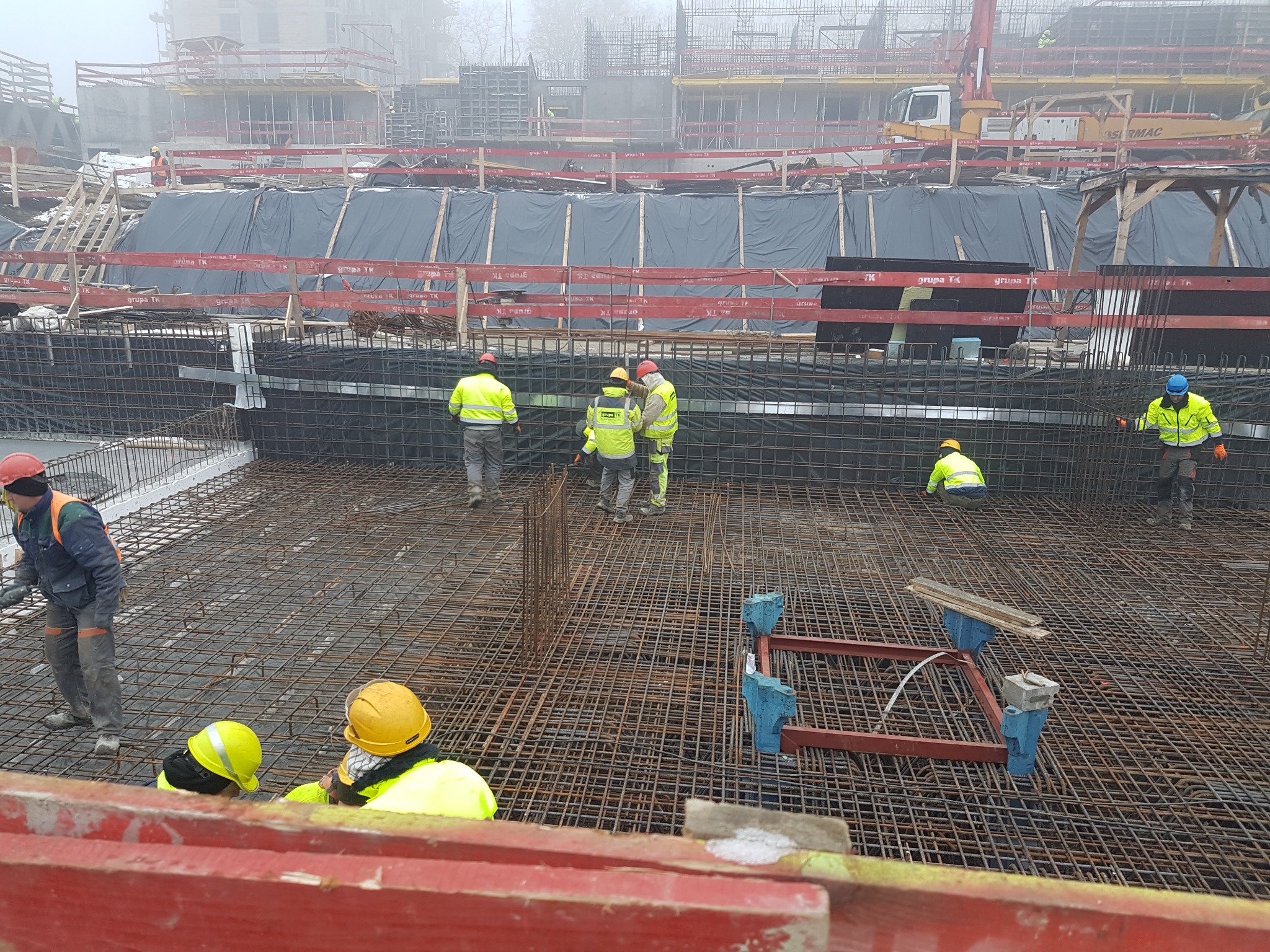 Ekipa budowlana w jaskrawych kamizelkach pracuje nad konstrukcją z siatki stalowej na placu budowy w mglisty dzień, widoczne szalunki i dźwig w tle.