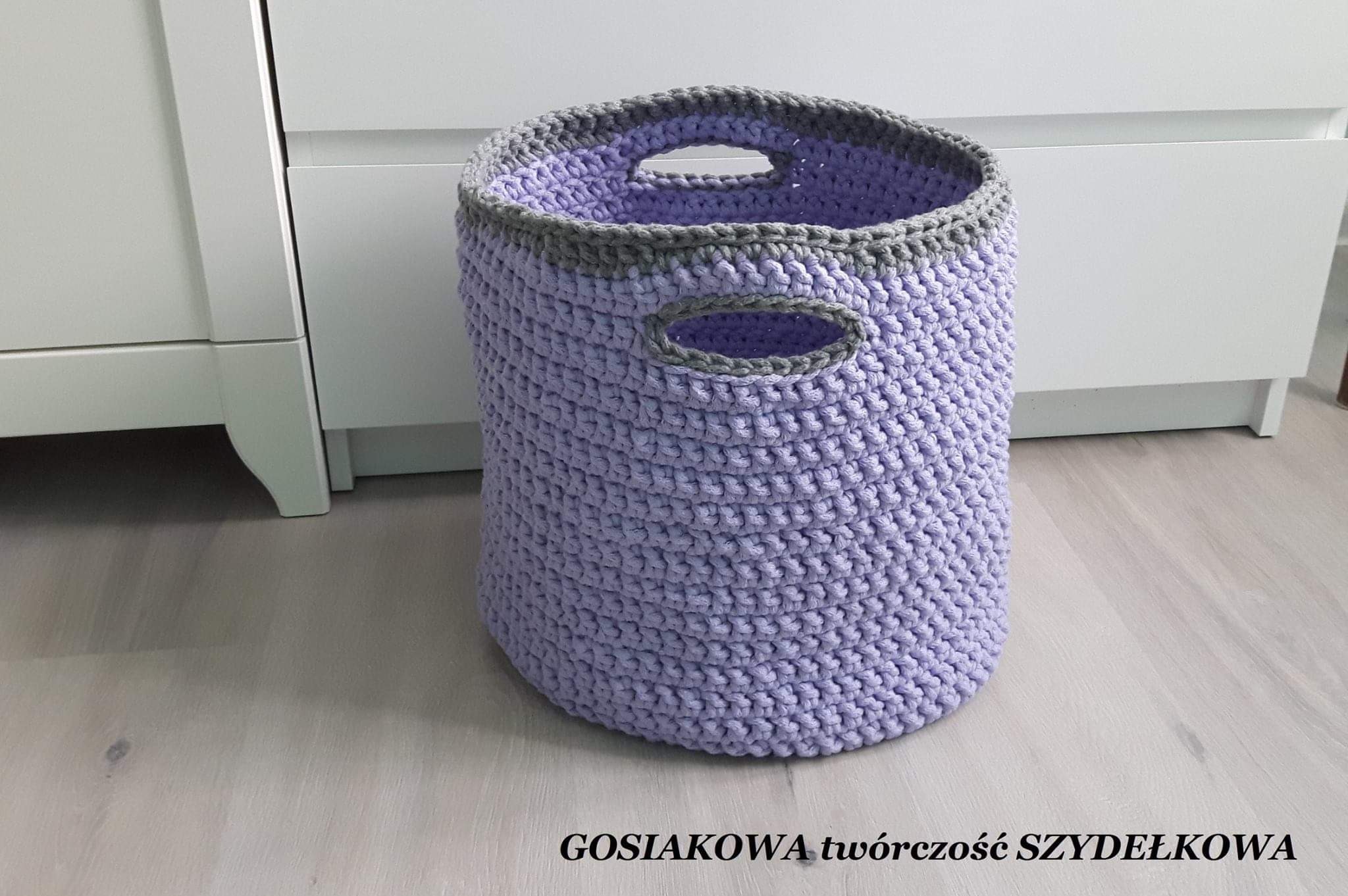 Ręcznie robiony, fioletowy kosz z uchwytami, wykonany na szydełku z grubej włóczki, z szarym wykończeniem, stojący na jasnej podłodze, obok białej komody.