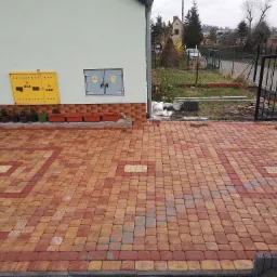 Ułożona kostka brukowa w odcieniach czerwieni i żółci, z widocznymi elementami infrastruktury budynku, takimi jak skrzynka gazowa i przyłącze elektryczne, oraz ogrodzeniem w tle.