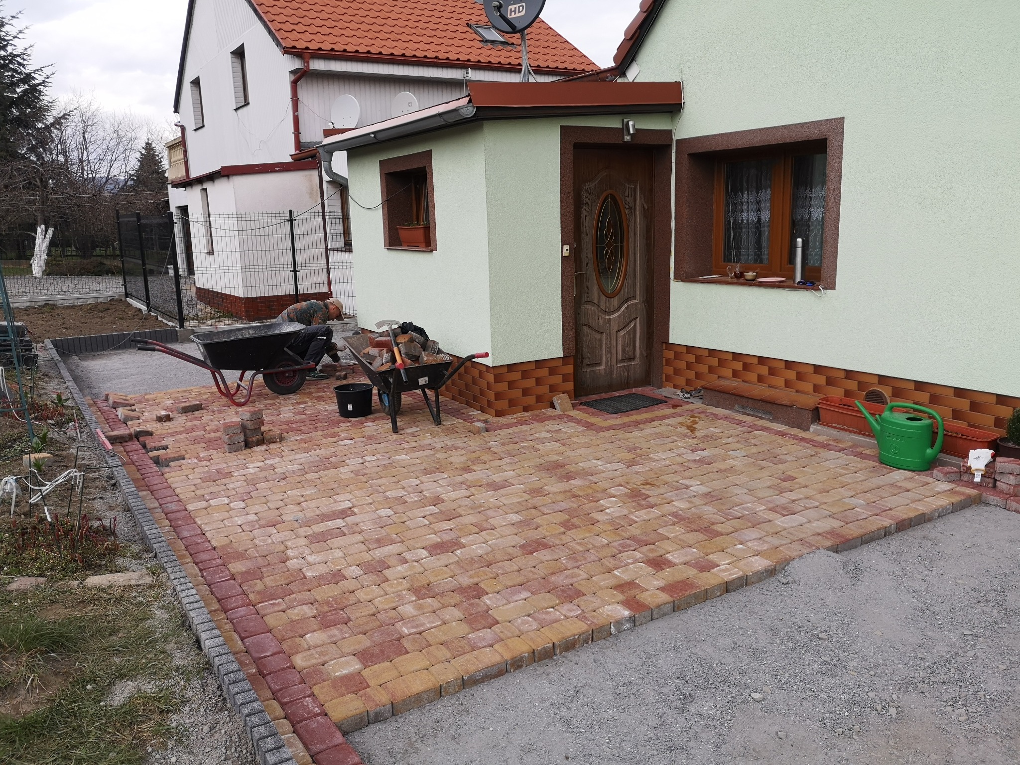 Układanie kostki brukowej w odcieniach czerwieni i beżu na posesji przed domem, widoczny pracownik z taczką, etap prac wykończeniowych.
