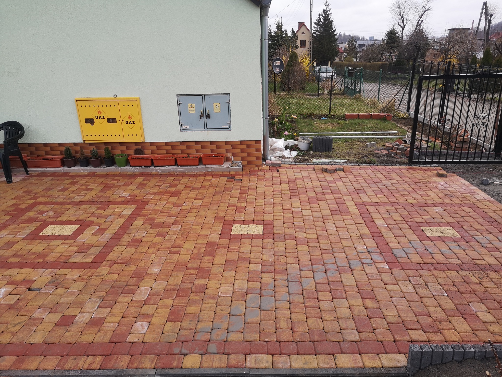 Ułożona kostka brukowa w odcieniach czerwieni i żółci, z widocznymi elementami infrastruktury budynku, takimi jak skrzynka gazowa i przyłącze elektryczne, oraz ogrodzeniem w tle.