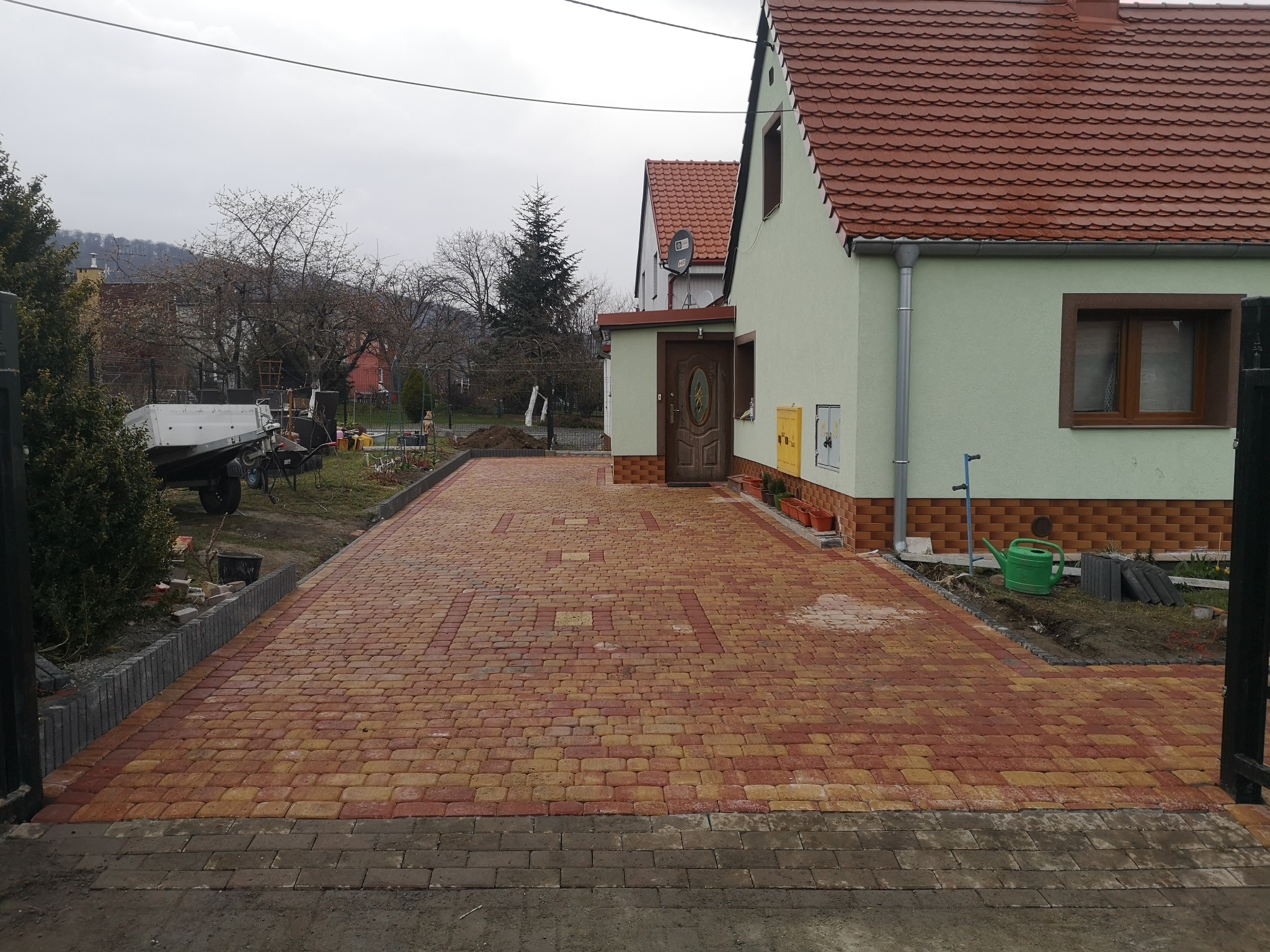 Nowo ułożona, dwukolorowa kostka brukowa w odcieniach czerwieni i żółci na podjeździe przed domem z czerwonym dachem, widok zza częściowo otwartej bramy w pochmurny dzień.
