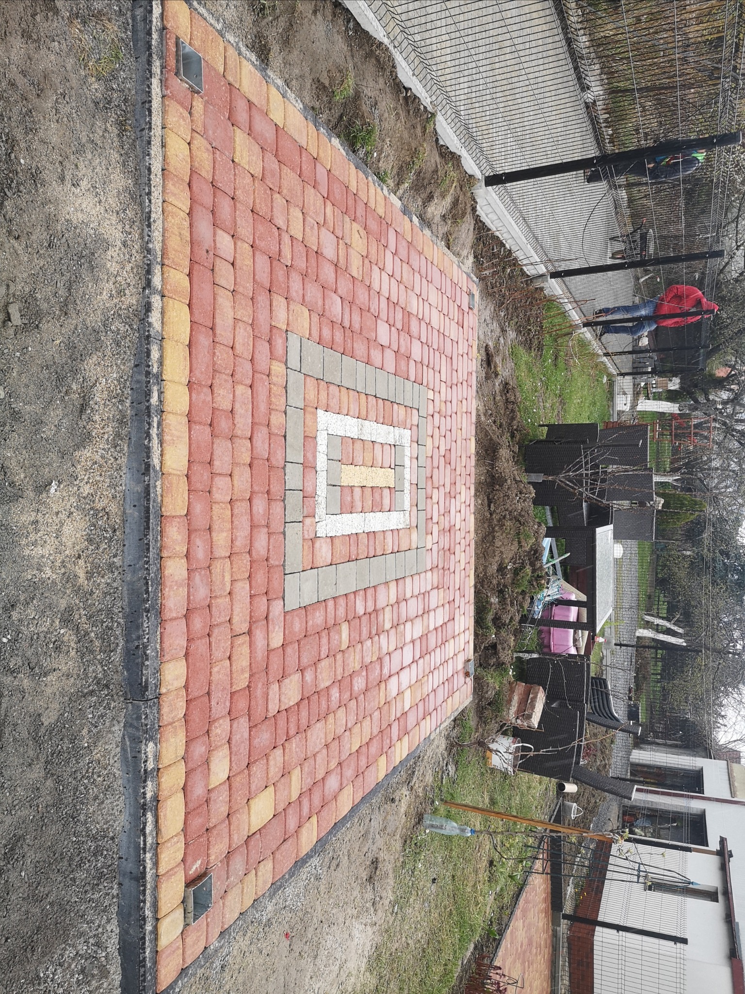 Świeżo ułożona kostka brukowa w odcieniach czerwieni i żółci z centralnym wzorem kwadratu w kolorze szarym i białym, widok z góry na tle ziemi i ogrodzenia.