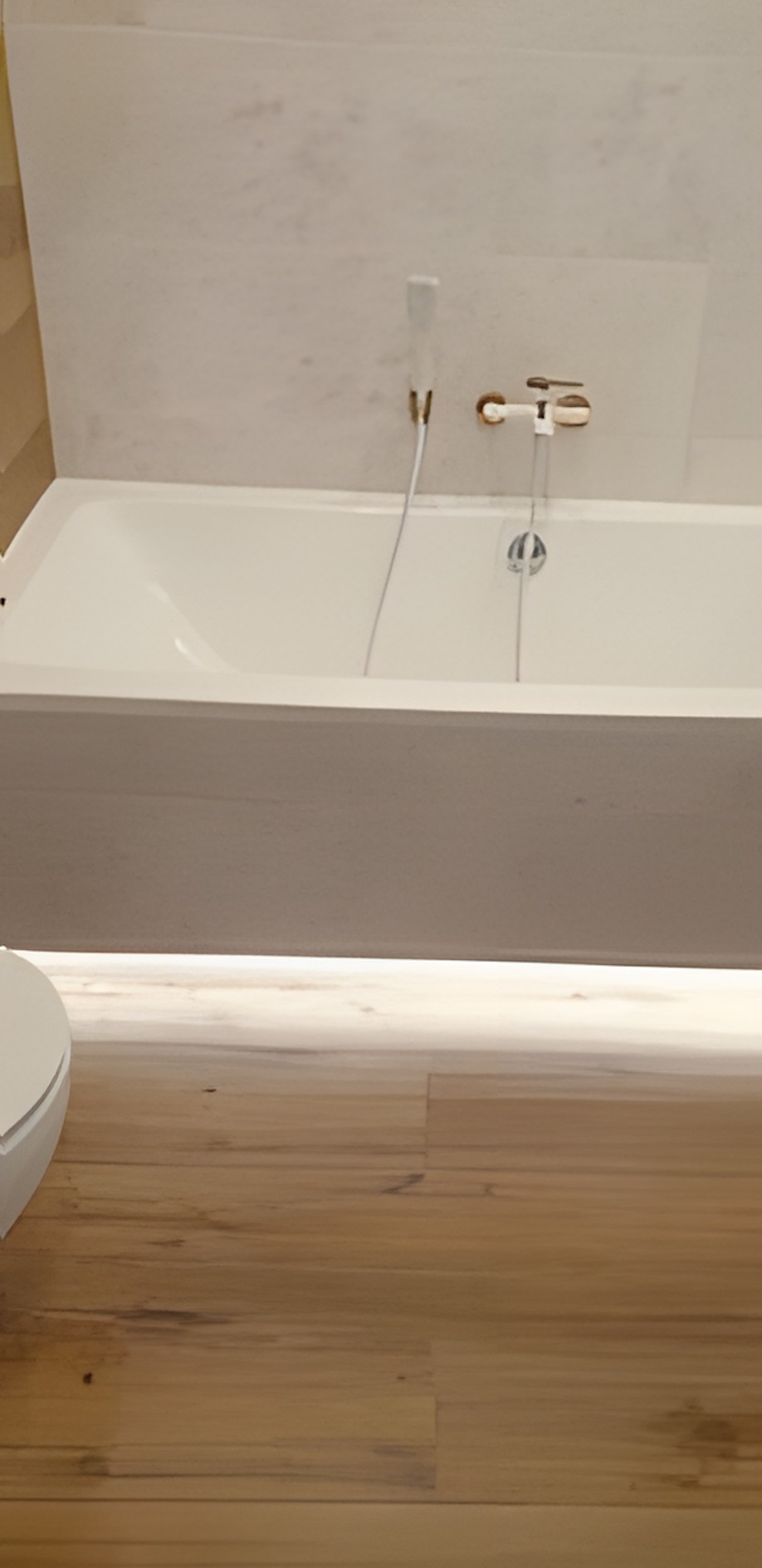 Nowoczesna łazienka z białą wanną, armaturą w odcieniach złota i podświetlaną podłogą z drewnopodobnych paneli. Minimalistyczny design.