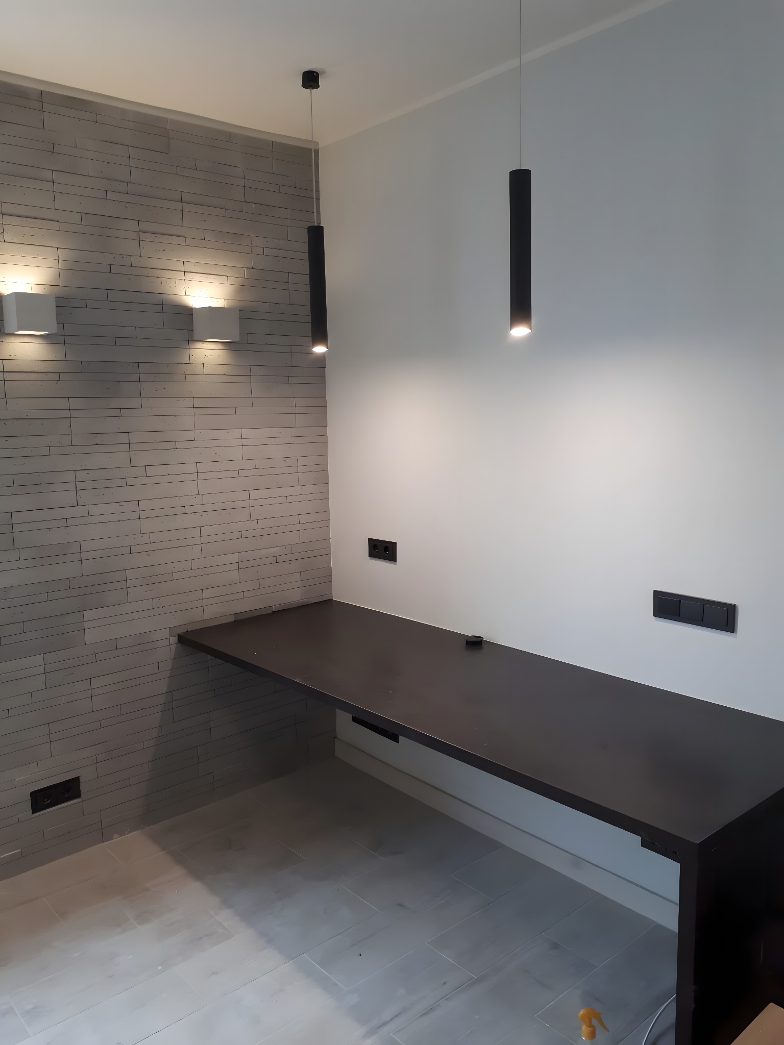 Minimalistyczne wnętrze: szara ściana z cegły, długa, czarna półka, podwieszane lampy cylindryczne i gniazdka elektryczne. Nowoczesny design, neutralna kolorystyka.