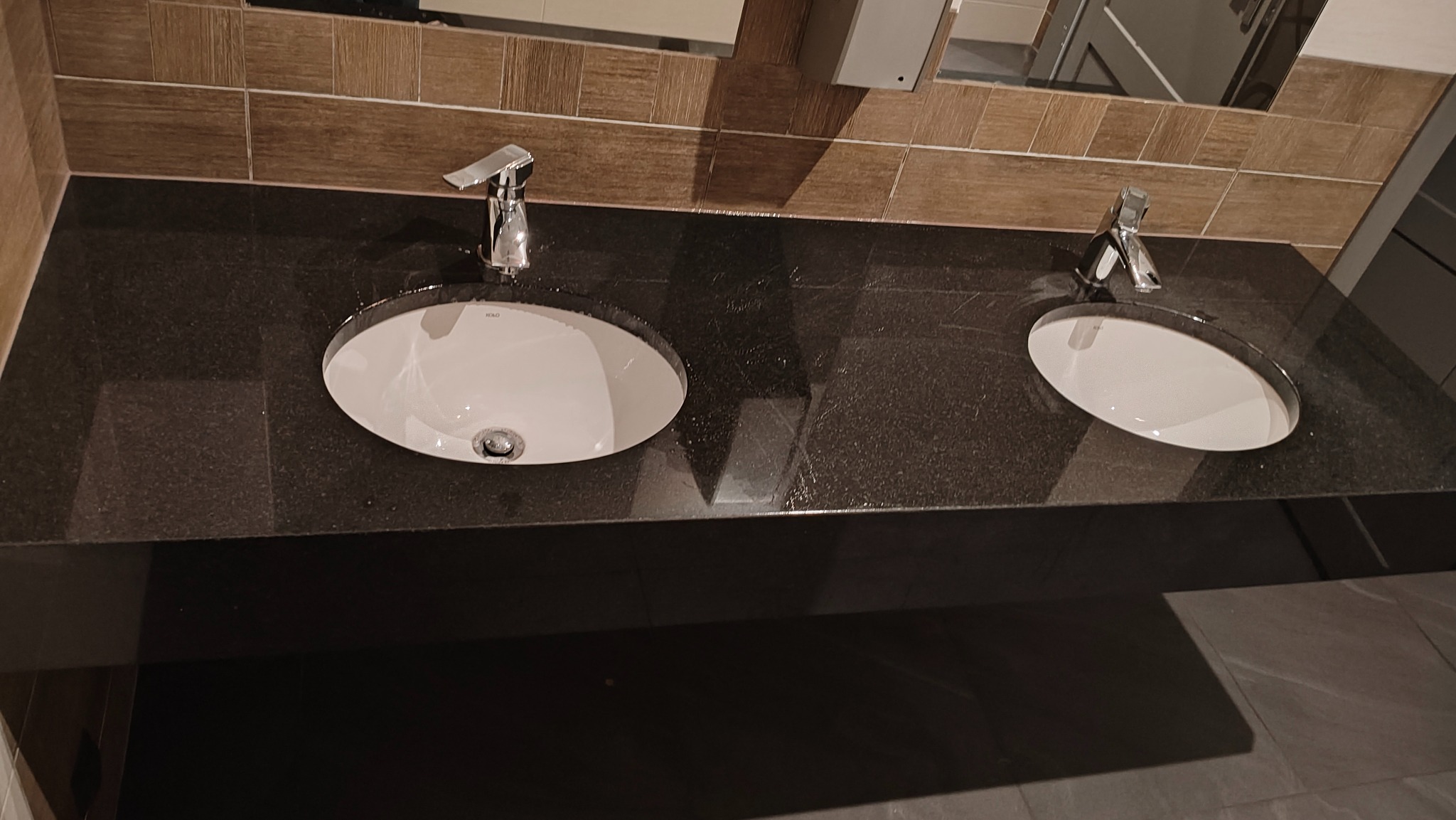 Dwie umywalki ceramiczne wpuszczone w czarny, błyszczący blat. Nad nimi lustro i brązowe płytki imitujące drewno. Nowoczesna armatura.