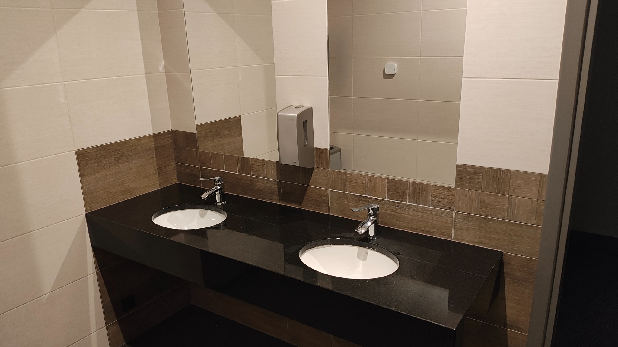 Nowoczesna łazienka z dwoma umywalkami na czarnym blacie, jasne i brązowe płytki na ścianach, duże lustro, dozownik mydła. Ukończona realizacja.