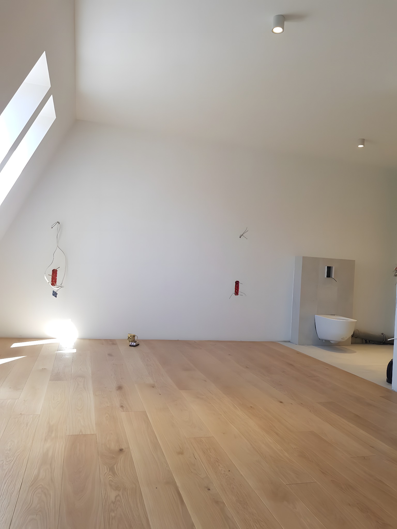 Jasna, drewniana podłoga w nowym, jasnym pokoju na poddaszu z oknem dachowym i instalacją elektryczną na ścianie. Widoczny fragment toalety.