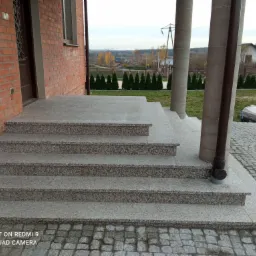 Granitowe schody wejściowe do domu z cegły, prowadzące na taras wyłożony kostką brukową, z widokiem na oddalony krajobraz.