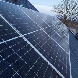 Szel Klima - Panele fotowoltaiczne na dachu domu jednorodzinnego, widok z dołu, błękitne niebo w tle. Widoczny fragment dachu i komin.