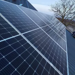 Panele fotowoltaiczne na dachu domu jednorodzinnego, widok z dołu, błękitne niebo w tle. Widoczny fragment dachu i komin.