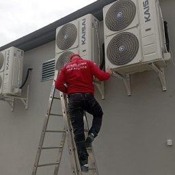 Szel Klima - Instalator na drabinie montuje klimatyzatory marki Kaisai na szarej ścianie budynku. Widoczne trzy jednostki zewnętrzne, montaż na wspornikach.