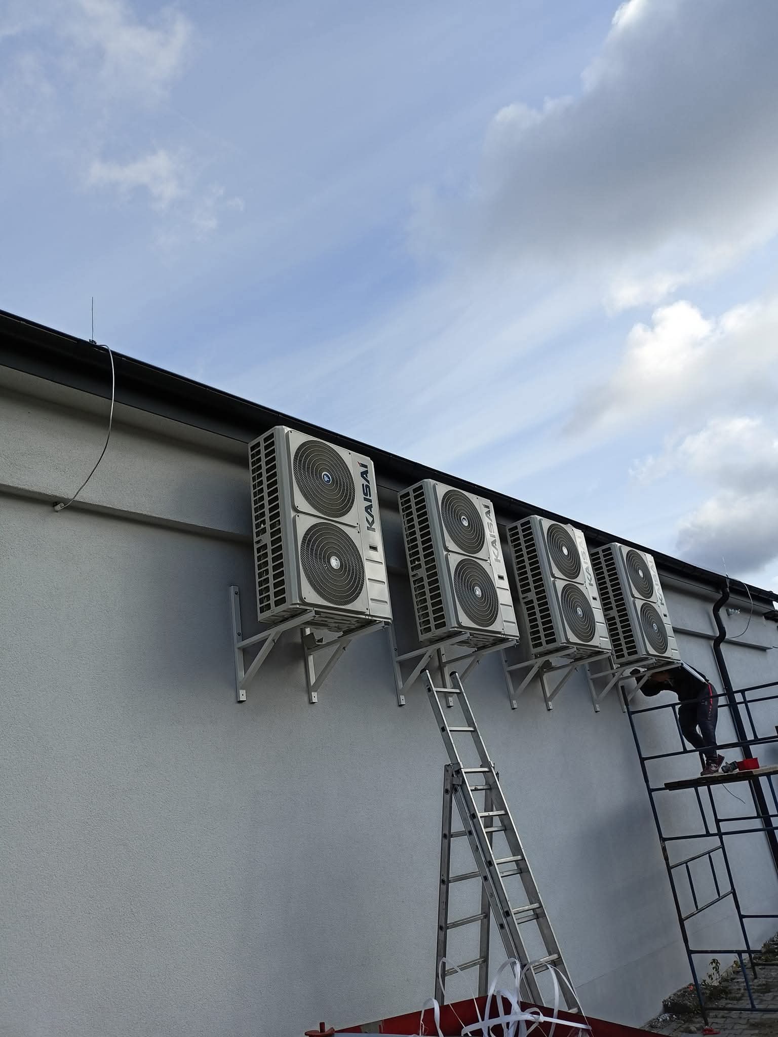 Montaż klimatyzacji Kaisai na elewacji budynku. Cztery jednostki zewnętrzne zamontowane szeregowo. Pracownik na rusztowaniu podczas instalacji. Błękitne niebo z chmurami w tle.