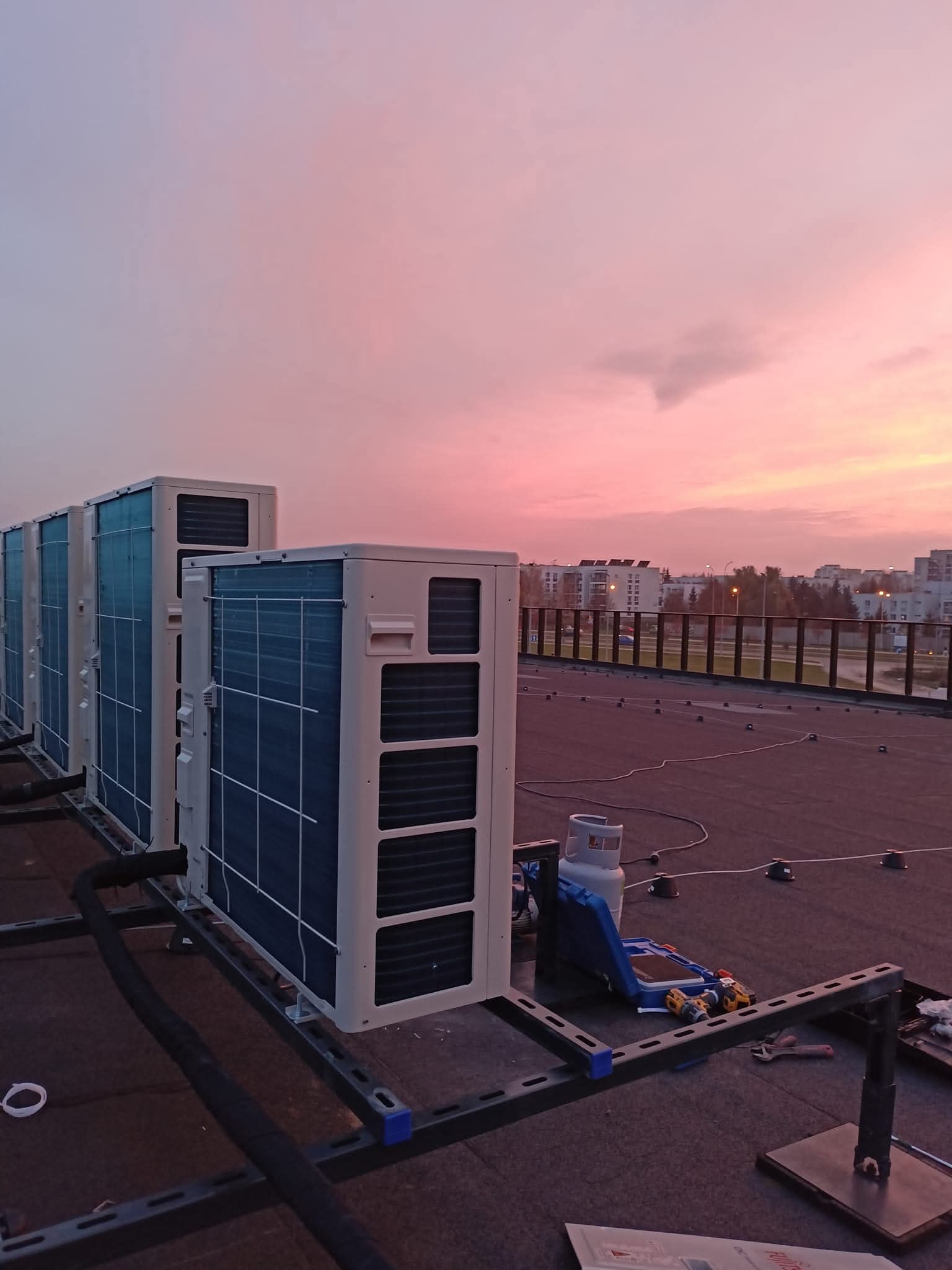 Rząd klimatyzatorów na dachu budynku o zmierzchu. Widoczne narzędzia montażowe i elementy instalacji. W tle zabudowa miejska podświetlona światłami ulicznymi.