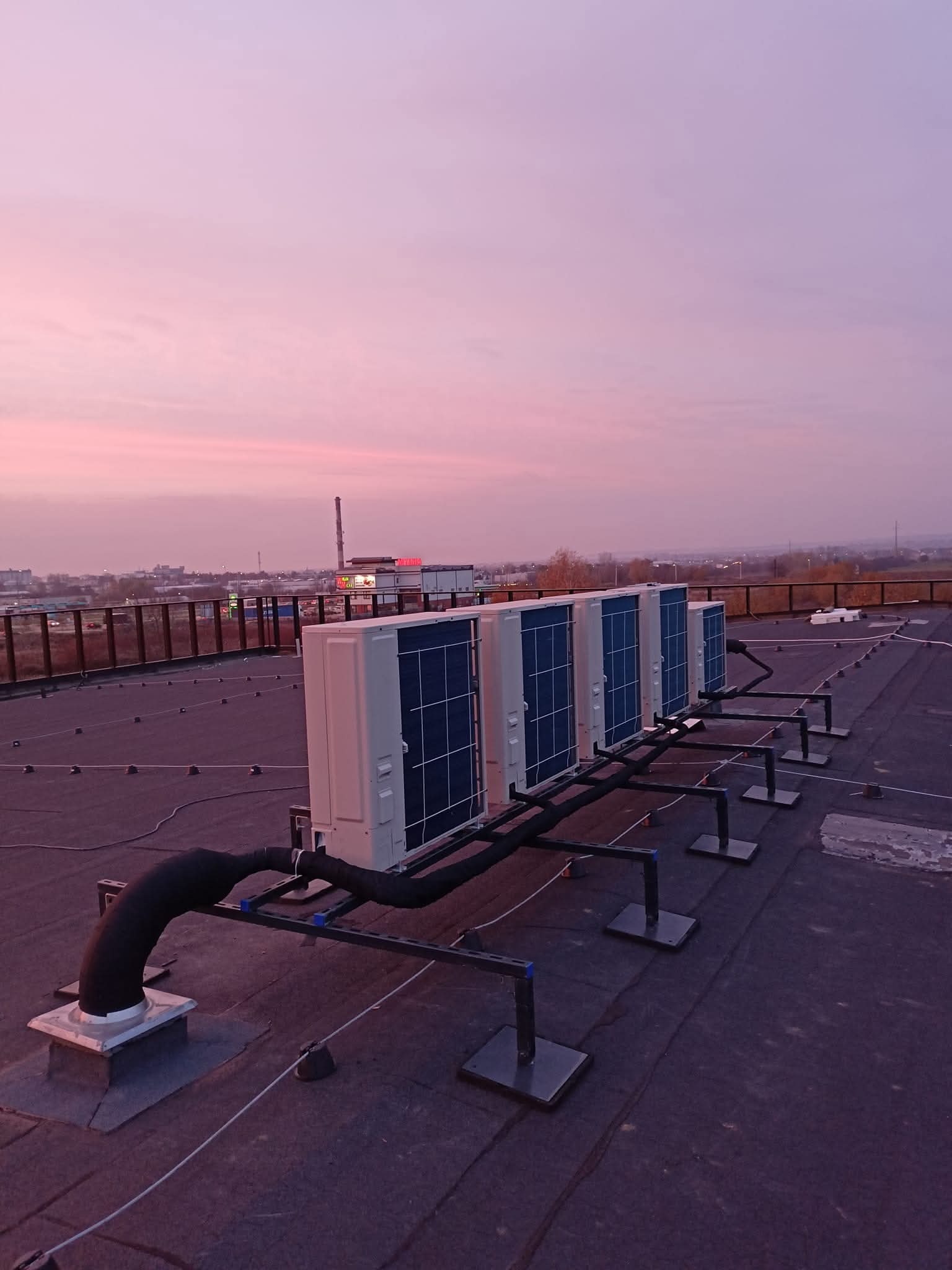 Rząd klimatyzatorów na dachu budynku komercyjnego, z widokiem na miasto o zmierzchu. Cztery jednostki zewnętrzne zamontowane na czarnych wspornikach.