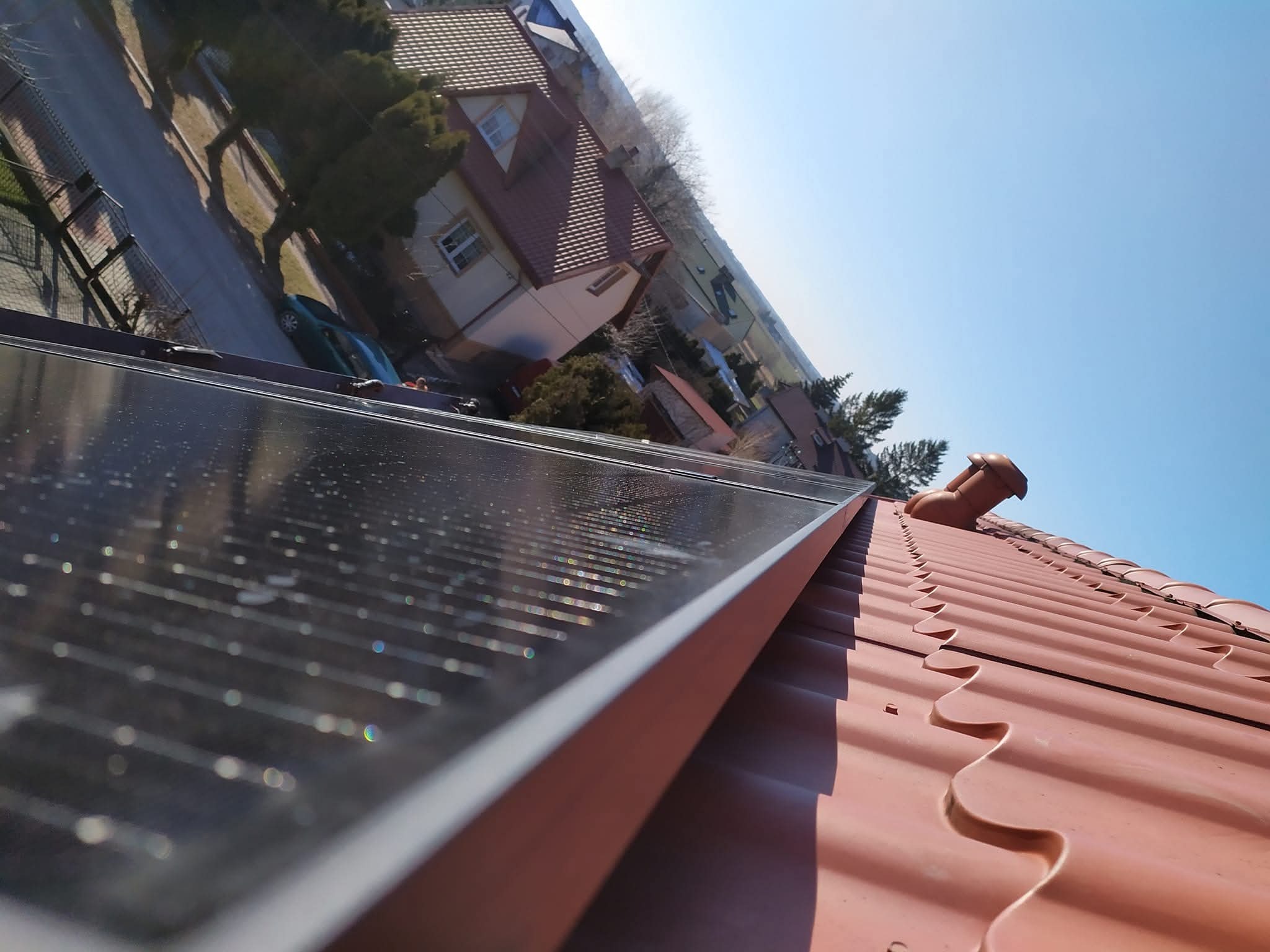 Panel fotowoltaiczny, widok z góry na czerwony dach domu w słoneczny dzień. Na panelu widoczne odbicia otoczenia. Ujęcie z niskiego kąta.