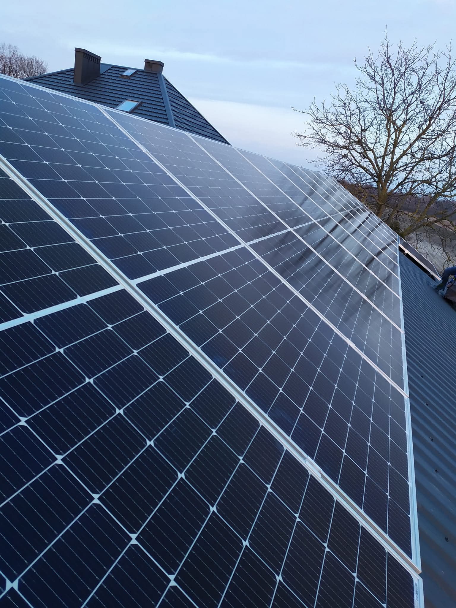 Panele fotowoltaiczne na dachu domu jednorodzinnego, widok z dołu, błękitne niebo w tle. Widoczny fragment dachu i komin.