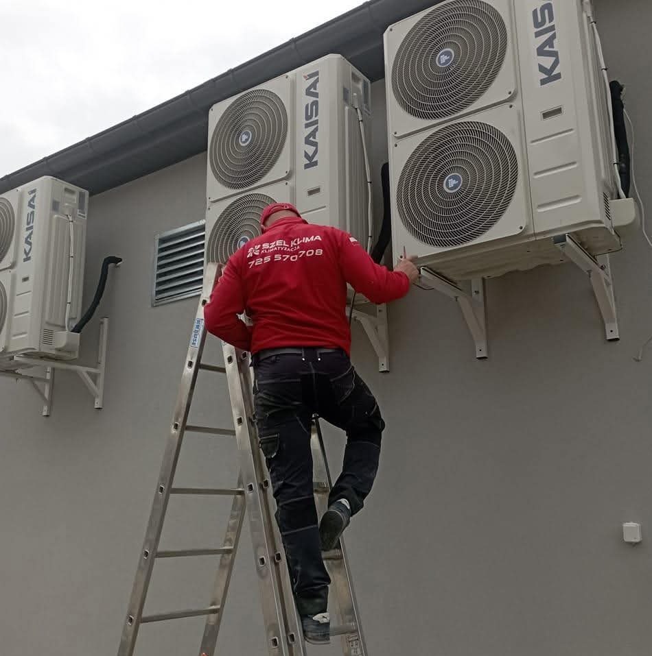 Instalator na drabinie montuje klimatyzatory marki Kaisai na szarej ścianie budynku. Widoczne trzy jednostki zewnętrzne, montaż na wspornikach.