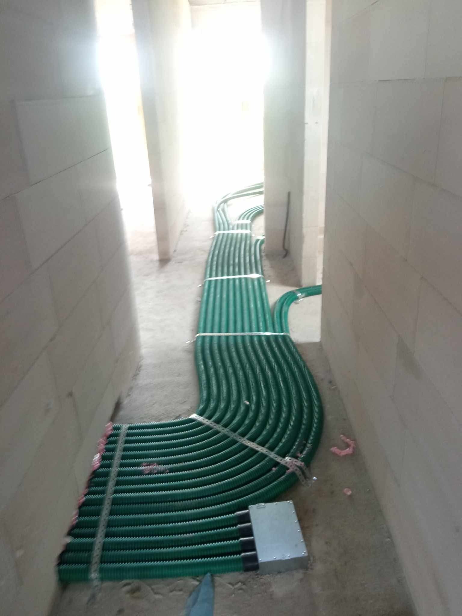 Zielone rury wentylacyjne ułożone równolegle w korytarzu budynku w trakcie budowy, przymocowane metalową taśmą do ściany, z widoczną puszką rozdzielczą.