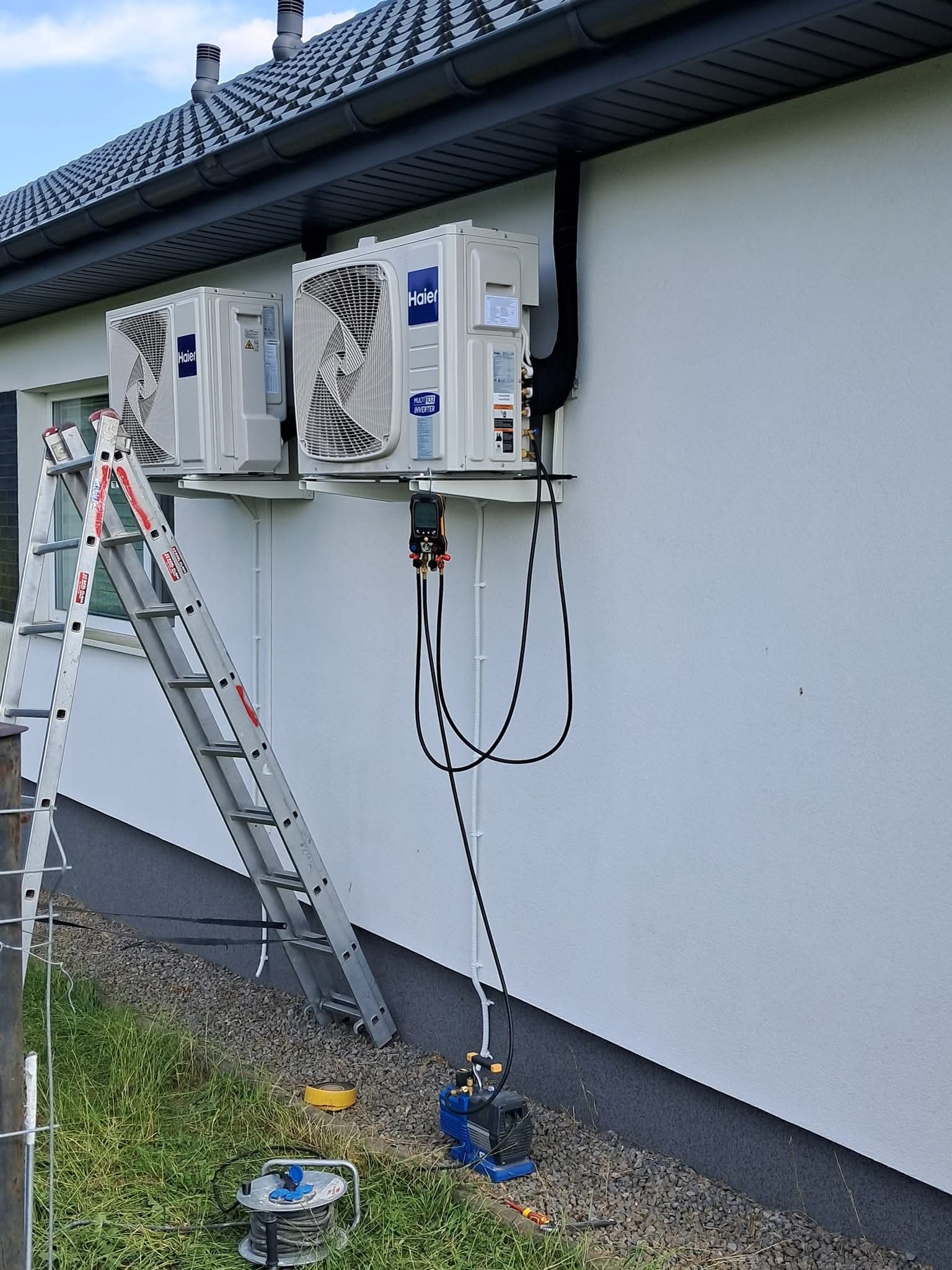 Dwa klimatyzatory Haier zamontowane na elewacji domu. Widoczna drabina, przewody instalacyjne i narzędzia, co sugeruje etap montażu lub serwisu klimatyzacji.