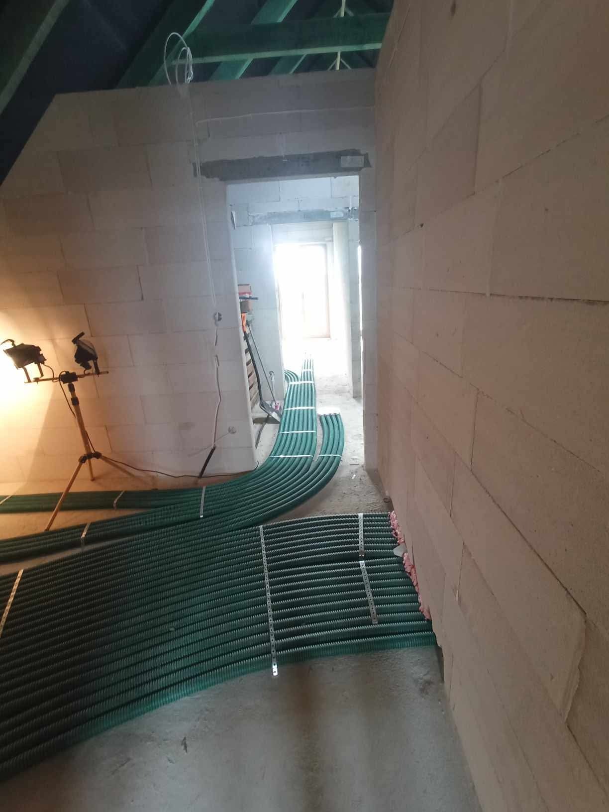 Wnętrze w trakcie prac: rury wentylacyjne ułożone wzdłuż ściany z betonu komórkowego, oświetlone lampą budowlaną. Widoczny surowy stan wykończenia.
