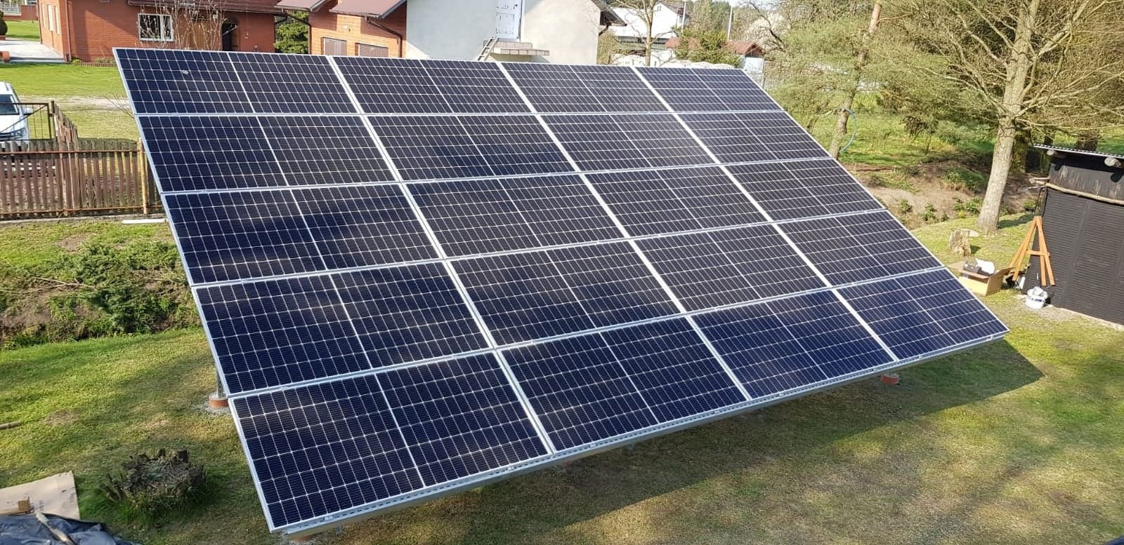 Zestaw paneli słonecznych zamontowany na metalowej konstrukcji na trawniku przed domem jednorodzinnym, widoczne narzędzia i akcesoria montażowe obok paneli.