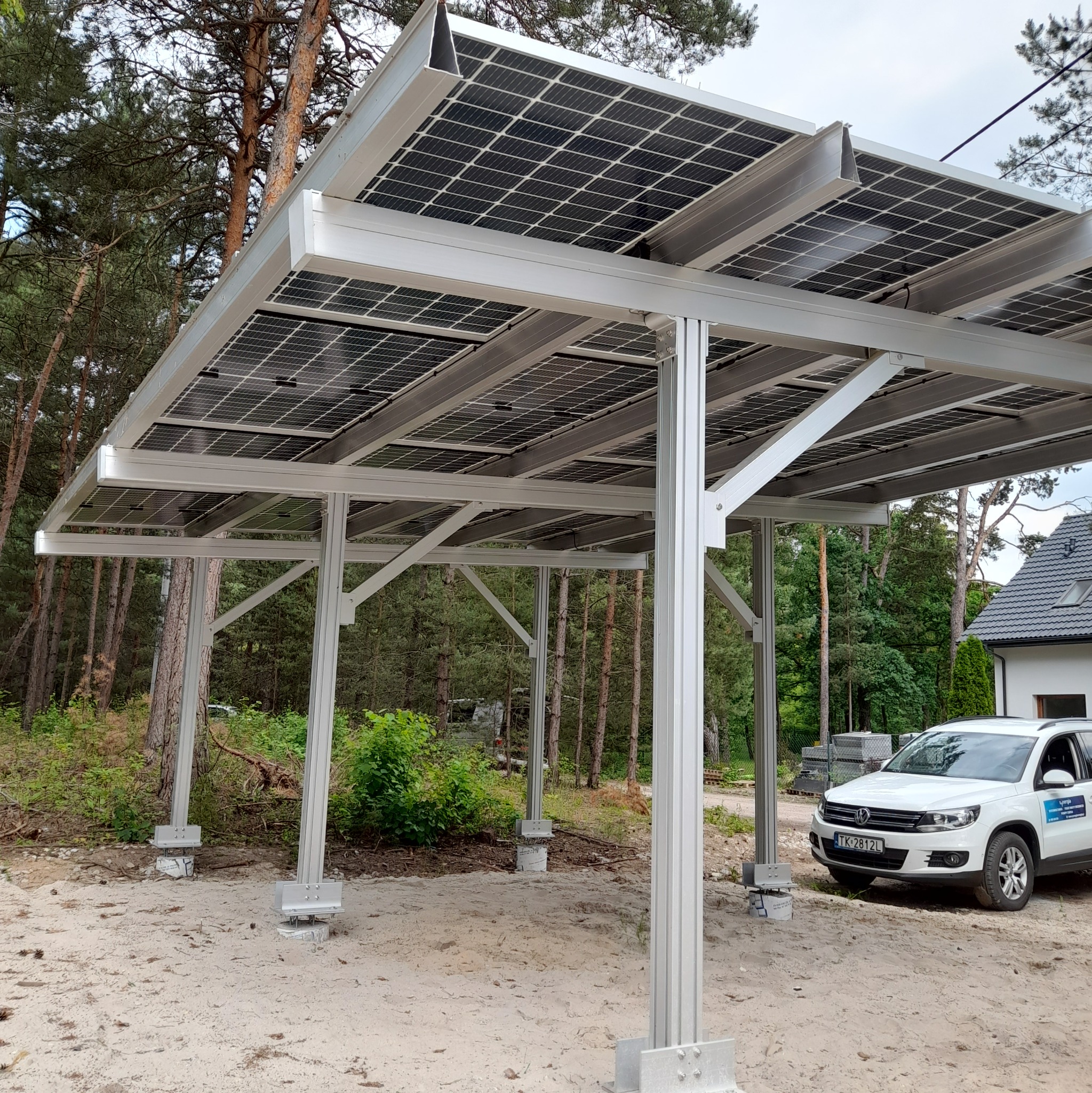 Zadaszenie z paneli słonecznych na aluminiowej konstrukcji, stojące obok białego samochodu na piaszczystym podłożu z widocznymi drzewami w tle.