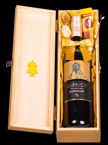 Otwarty drewniany kosz prezentowy z butelką wina Zafrico, słodyczami, jajkiem przepiórczym, szyszką i gałązkami wierzby, przewiązany żółtą wstążką, z dekoracją w kształcie żółtego kurczaczka na boku.