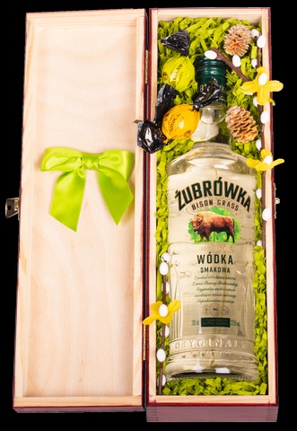 Drewniana skrzynka prezentowa z butelką Żubrówki, cukierkami w czarnych i żółtych papierkach, wypełniona zielonym siankiem i ozdobiona gałązkami z małymi szyszkami i żółtymi kwiatkami, zamknięta...
