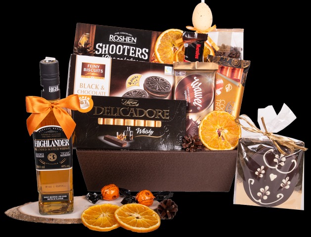 Elegancki kosz prezentowy z whisky, czekoladkami, ciastkami i suszonymi pomarańczami, ułożony na drewnianej podstawce.