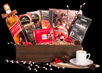 Zestaw prezentowy w drewnianej skrzynce z butelką whisky Red Label, kawą Lavazza, czekoladkami Ritter Sport i innymi słodyczami, udekorowany szyszkami i gałązkami, z filiżanką espresso obok.