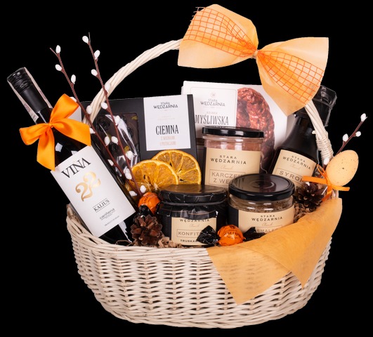 Biały wiklinowy kosz prezentowy wypełniony winem, słodyczami, konfiturami, syropem i innymi produktami spożywczymi, ozdobiony pomarańczową wstążką i gałązkami wierzby.
