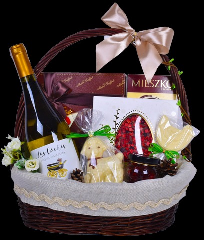 Elegancki wiklinowy kosz wypełniony winem, czekoladkami, konfiturą, figurką baranka w folii i kartką z grafiką lokomotywy, ozdobiony beżową kokardą i koronkową wstążką.