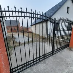 Czarna, metalowa brama wjazdowa z dekoracyjnymi grotami, zamontowana między pomarańczowymi, betonowymi słupkami, na tle białego domu z ciemnym dachem i ceglanego budynku.