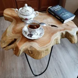 Stolik kawowy z blatem z litego drewna o nieregularnym kształcie, wsparty na czarnych, metalowych nogach, z filiżanką herbaty, cukierniczką i książką na blacie.