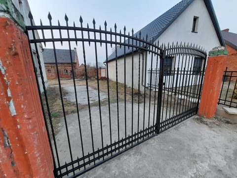 Czarna, metalowa brama wjazdowa z dekoracyjnymi grotami, zamontowana między pomarańczowymi, betonowymi słupkami, na tle białego domu z ciemnym dachem i ceglanego budynku.