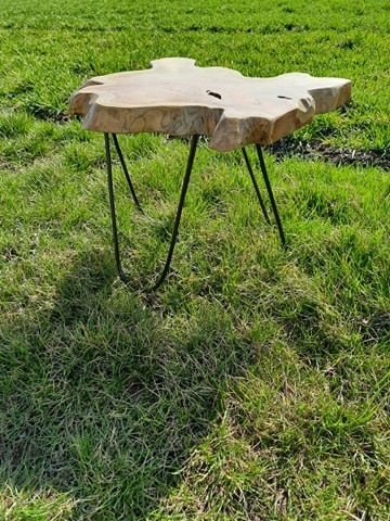 Niski stolik kawowy z blatem z surowego drewna o nieregularnym kształcie, wsparty na trzech czarnych, metalowych nogach w stylu hairpin, ustawiony na trawniku.