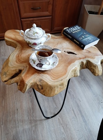 Stolik kawowy z blatem z litego drewna o nieregularnym kształcie, wsparty na czarnych, metalowych nogach, z filiżanką herbaty, cukierniczką i książką na blacie.